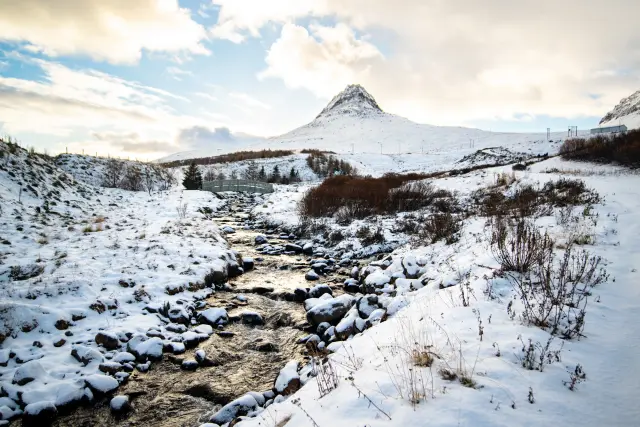 A snow-covered stream and small bridge below a sharp peak, Iceland