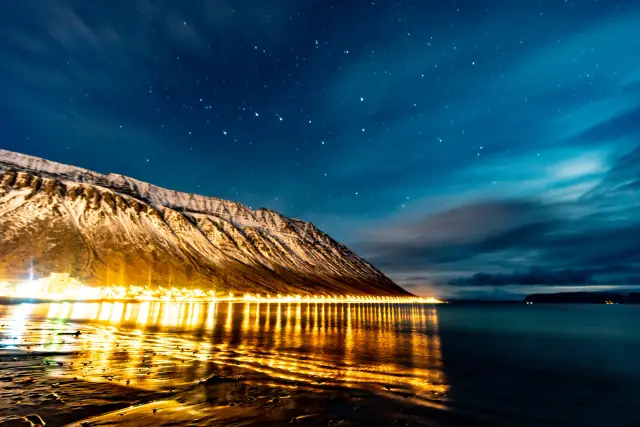 Golden town lights reflected in a fjord beneath a snowy mountain and starry sky, Iceland