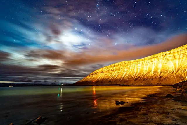 A long-exposure night scene of a starry sky over a calm bay with a warmly lit coastal cliff.