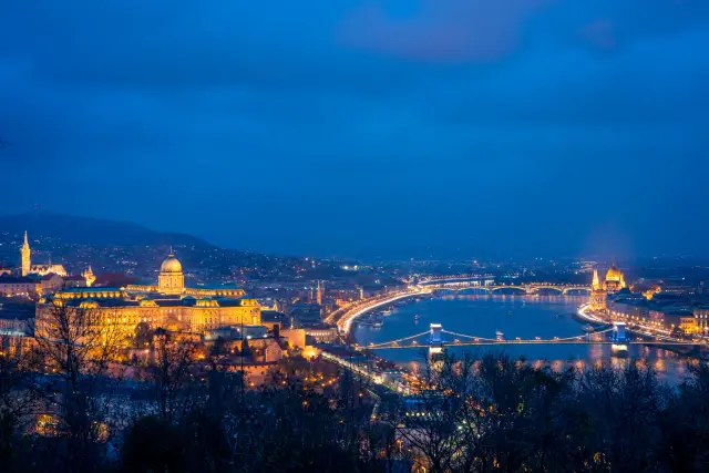 An elevated view of Buda Castle, the Chain Bridge and the Danube illuminated at blue hour.